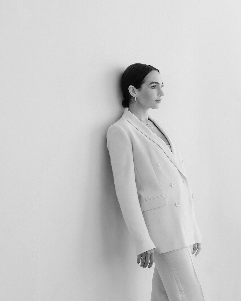 A black and white photo of pianist Emily Bear leaning against a wall in a white suit.