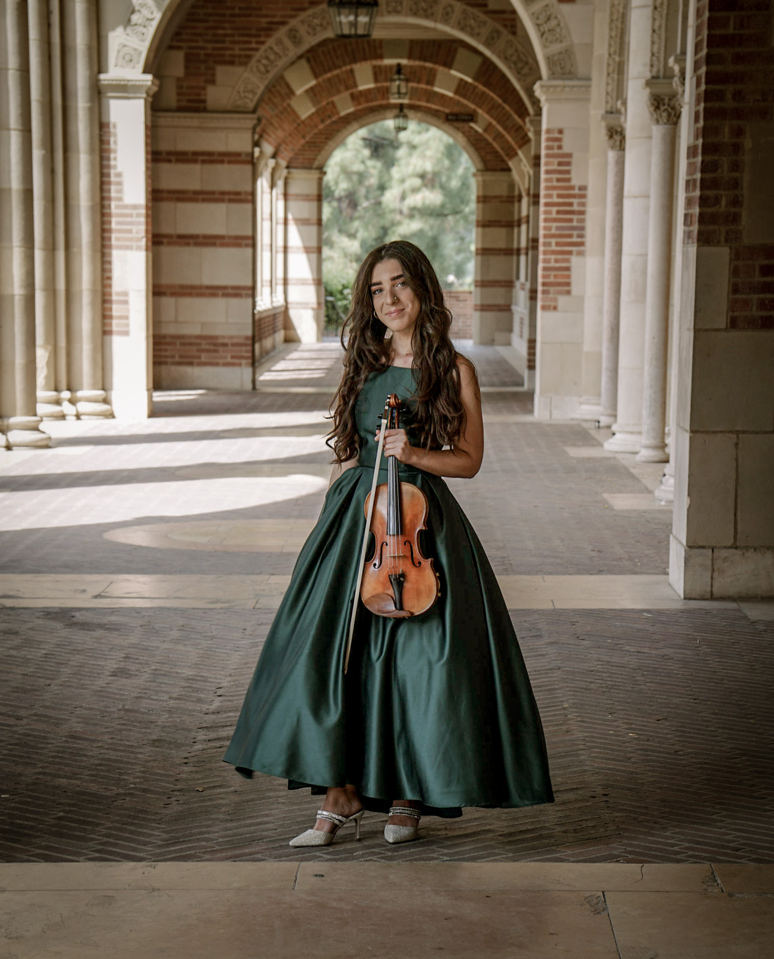 A picture of new LAJS intern Sarah Seltzer, standing in an archway in a green dress while holding her violin.