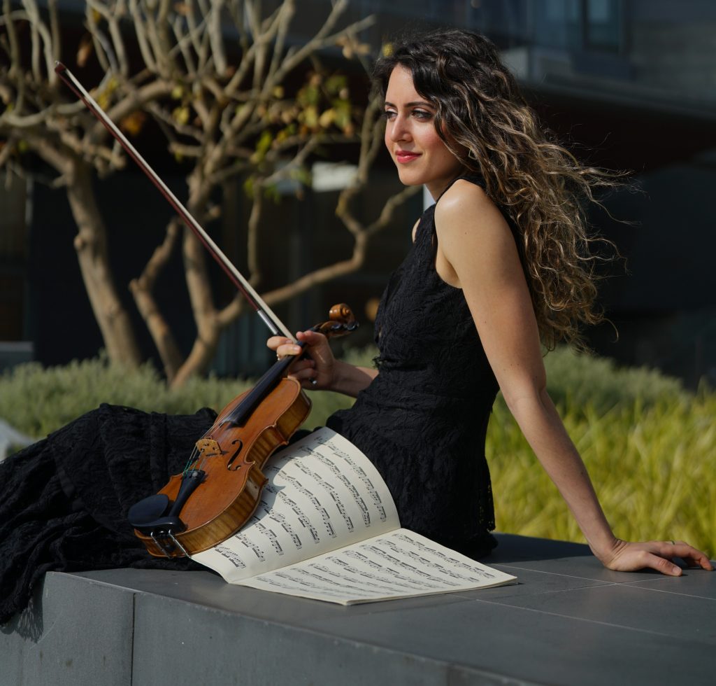 Headshot of Chloe Pourmorady sitting outside with sheet music and her violin